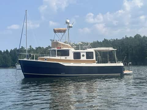 Cruising trawler at anchor in a quiet pine-lined cove with a sailboat in the background.